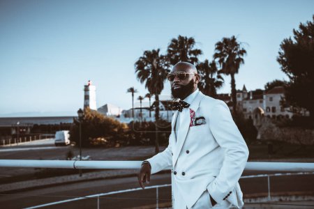 Photo for An irresistible bald, bearded black businessman, in a fashionable suit, sunglasses, and a polka dot bow tie standing against a metal fence on a terrace with palm trees and a lighthouse in the back - Royalty Free Image
