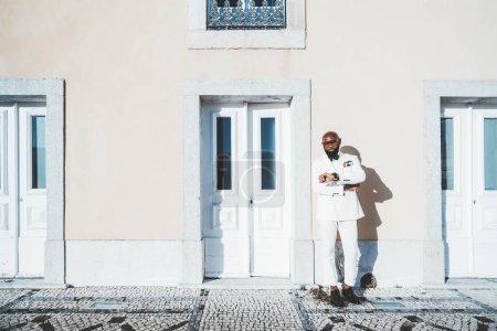 Photo for An African-American, well-groomed, bearded male wearing a white suit leaning against the pastel wall of a classic building with tall doors. The businessman is checking his wristwatch on the sidewalk - Royalty Free Image