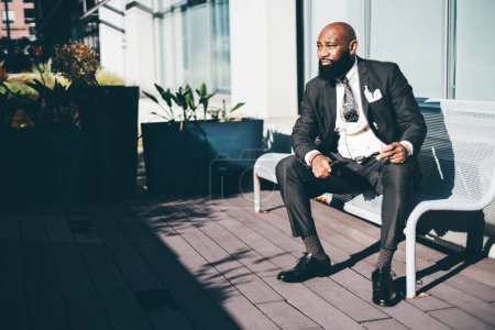 Photo for An elegant unshaved bald African businessman in a sophisticated tailored suit with a polka-dot tie, holding eyeglasses and a phone sitting on a bench on a veranda with plants in planters under the sun - Royalty Free Image