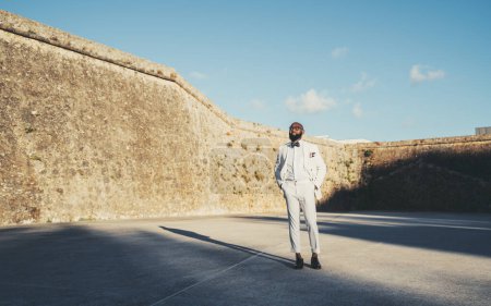 Photo for An African bald and bearded man well-dressed in a white suit with a bow tie, glasses, and leather shoes stands on the concrete floor; Surrounding him is the old wall of the village of Cascais - Royalty Free Image