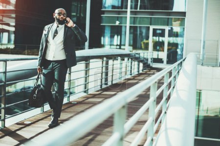 Photo for A white-collar worker carries a weekender while having a phone call when leaving the office. A hairless black man wearing sunglasses, a suit, a tie, and a white vest, exiting a contemporary building - Royalty Free Image