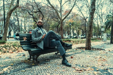 Foto de Un hombre negro calvo barbudo maduro y guapo emprendedor con un traje elegante sentado en un banco de madera en el pavimento; un hombre de negocios afroamericano que contempla la escena urbana en invierno - Imagen libre de derechos