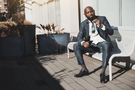 Photo for A sophisticated bald African businessman in a sophisticated tailored suit with a polka-dot tie, holding eyeglasses and a phone sitting on a bench on a terrace with plants in planters under the sun - Royalty Free Image