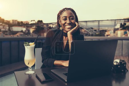 Foto de A portrait of a young cheerful African American woman entrepreneur using her laptop while sitting in an outdoor restaurant; a dazzling smiling black female freelancer in a street cafe with a netbook - Imagen libre de derechos