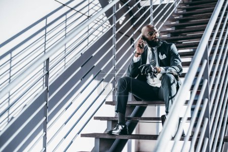 Foto de A serious fancy bald bearded black man entrepreneur in eyeglasses, a tailored formal suit, and leather gloves is sitting on a step of an outdoor staircase and talking on the phone while looking aside - Imagen libre de derechos