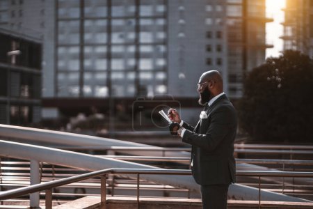 Téléchargez les photos : A side view of a silhouette of a bald fancy bearded black man entrepreneur taking notes in documents while standing on the street; an African American businessman outdoors writing something on a paper - en image libre de droit