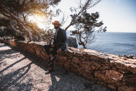Téléchargez les photos : A wide-angle shot of a sophisticated mature bearded black guy in a straw hat and a tailored elegant summer suit is sitting on a stone fence of a coastal park with a seascape and sunset behind him - en image libre de droit