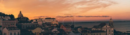 Foto de A panoramic image with a cityscape of an antique part of Lisbon with old houses, churches, and a river with a stunning sunset in the background; a panorama of an old Portuguese city, dramatic evening - Imagen libre de derechos