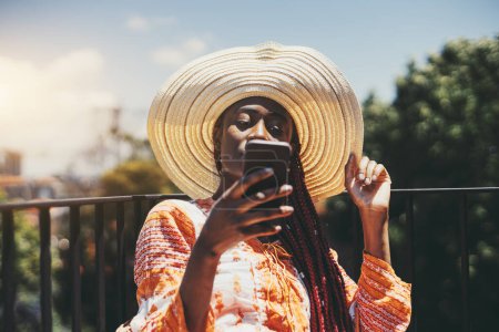 Téléchargez les photos : A portrait of a ravishing young black female in a summer dress and a hat with a wide brim phoning while sitting on her balcony on a warm day; African American woman in a sundress and a shovel hat - en image libre de droit