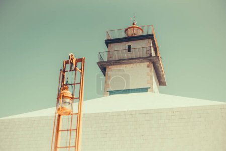 Téléchargez les photos : A wide low-angle shot of an old white-colored lighthouse with a horizontal line as detail made with tiles, and in front of it an orange ladder with a container attached to it, under the blue sky - en image libre de droit