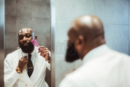 Téléchargez les photos : A capture of a mature well dressed fully bearded bald black male in a white vest with a polka-dot necktie combing his beard with a pink comb in front of a narrow mirror, in a grey-stoned restroom - en image libre de droit