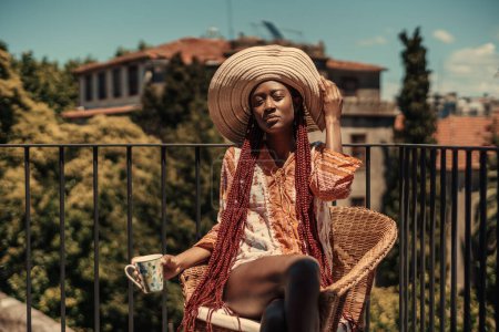 Foto de A colorful capture of a young girl of Afro-descendant origin, dressed femininely, in a pink and orange flowery dress, wide-brimmed hat, and long hair, on an rooftop while soaking up the sun - Imagen libre de derechos