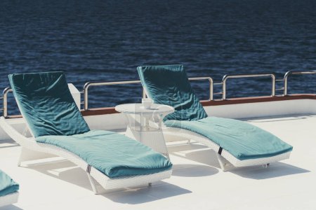 Téléchargez les photos : A wide-angle shot of two lounge chairs with blue cushions on top of them and a small white color coffee table on the upper deck of a luxurious sailing yacht on the sea, in the Maldives island - en image libre de droit