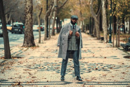 Téléchargez les photos : A dandy black man in a grey chequered vest and wool-blend coat, tailored trousers, and a beret, standing on the paving stone filled with orange leaves on a city alleyway surrounded by trees and cars - en image libre de droit