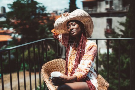Foto de A capture of a young feminine black girl, with long colorful braids, wearing a spring dress, a wide-brimmed hat that she's holding with one hand, and a mug in the other while soaking up some sun - Imagen libre de derechos
