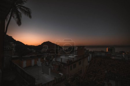 Photo for A wide-angle sunset shot from a rooftop in a house placed in a Brazilian favela in Rio, overlooking the calming sea and the rooftops of the surrounding houses with visible wear and tear of poverty - Royalty Free Image