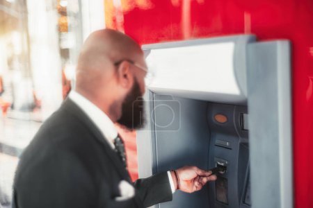 Téléchargez les photos : A shallow depth-of-field shot with a selective focus on an elegant hairless black man's hand, inserting a credit card into the red and white ATM machine to withdraw cash - en image libre de droit