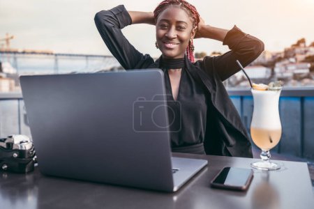 Foto de Portrait of a dazzling African freelancer using her laptop while sitting in an outdoor coffee, with a cocktail by her side, with her hands holding her long colorful hair while smiling for the camera - Imagen libre de derechos