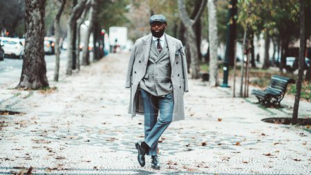 Photo for A mature African male with an unshaven black beard looking dapper in an elegant wool coat, beret, and suit. Standing in the middle of an autumn boulevard with his hands in the pockets - Royalty Free Image