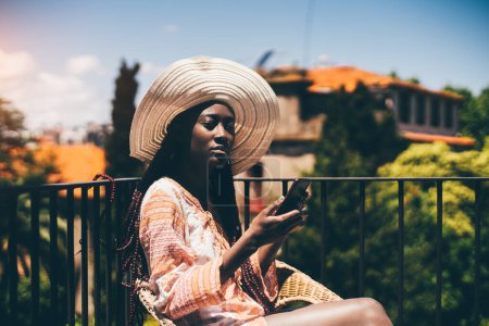 Foto de A shot of a young sensual woman with a wide-brimmed hat and long colorful braids, wearing a colorful oversized shirt, texting with both hands while soaking up some sun at the apartment balcony - Imagen libre de derechos