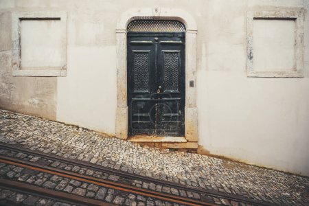 Téléchargez les photos : A black metal rusty door in a building with cemented windows, this worn-out building is losing its paint and it is situated on a sloping plane in a paved street where the tramway tracks can be seen - en image libre de droit