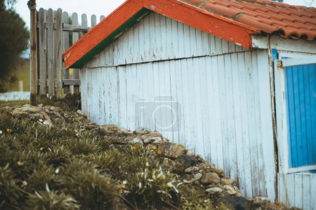 Téléchargez les photos : A capture of humble house built of wood painted white, and blue, but with orange tiles in an A shape situated on a small stone slope and undergrowth, the whole property is fenced in with wooden stakes - en image libre de droit