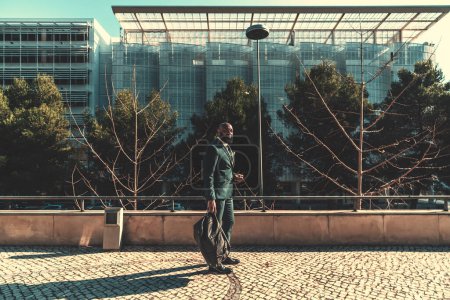 Foto de A sophisticated Angolan male was captured outside of the office building, in the back, standing on the pavement wearing a chequered dark green suit and carrying a weekender on a sunny day - Imagen libre de derechos