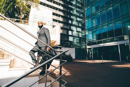 Téléchargez les photos : A black white-collar employee moving up the staircase, a metallic railing next to him, the office building with blue windows behind him and he is carrying a weekender bag, wearing dark grey suit - en image libre de droit