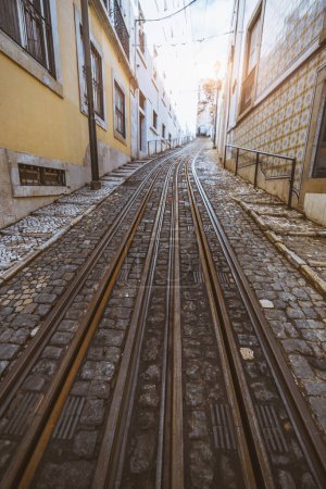 Photo for A vertical wide-angle view of a tramway lane over the Portuguese paving stone on a narrow picturesque Lisbon neighborhood in shades of yellow on the walls of the surrounding residential buildings - Royalty Free Image