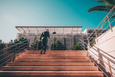 Photo for An African gallant in a full suit was captured from behind climbing up a large stairway toward a majestic and innovative glass-made office building exterior decorated with small bushes and trees - Royalty Free Image