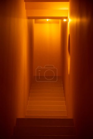 Foto de Vertical bottom-to-top shot of a building's interior stone staircase in a narrow and small hallway, illuminated with warm lighting, with shallow depth of field, and selective focus on middle steps - Imagen libre de derechos