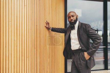 Photo for A gallant black man with drool and a bald head, wearing a tuxedo, black bow tie, and white shirt; Has one hand in his pocket and the other hand rests his body on a wall made of cherry wood stakes - Royalty Free Image