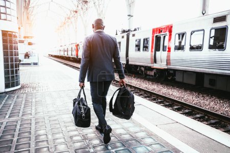 Photo for A backshot of a bald black elegant man well-dressed in a dark full-body suit, carrying a black backpack and a leather weekender, walking towards his train on the platform of the rail station - Royalty Free Image