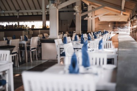 Foto de A capture using shallow depth of field and selective focus on the background of a restaurant that serves a resort, the white wooden dinner tables, already have silverware and blue napkins - Imagen libre de derechos