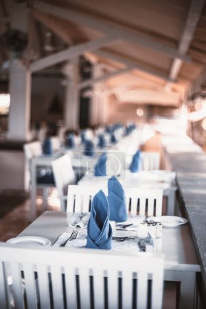 Téléchargez les photos : A selective focus on a white wooden dinner furniture table set, silverware, and blue napkins in a restaurant on a porch of a tropical resort in the pacific islands - en image libre de droit
