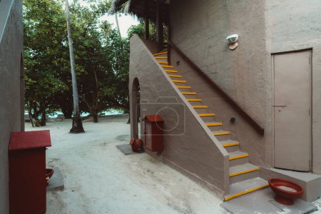 Téléchargez les photos : Tropical terracotta colored house villa housing entrance with sandy flooring, a red firefighting tool, and bowl for foot washing, yellow stripes highlighting the steps of the staircase - en image libre de droit