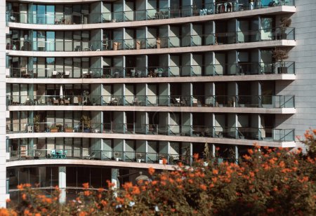 A glass-in facade of a tall white residential apartment building with balconies and windows in a pattern; a textured background of a block of flats in uptown on a bright day surrounded by flowers