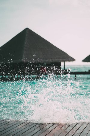 Photo for A vertical shot of the seawater splashing against the boardwalk in a defocused background, with a traditional stunning luxury overwater bungalow with canopy roofs on a Maldives island resort - Royalty Free Image