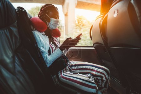 Téléchargez les photos : A young black girl dressed in a colorful vertical striped jumpsuit, white a facial mask, and a red neck pillow for the journey, texting while sitting on the black leather bus seat wearing - en image libre de droit