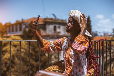 Photo for A young woman, with pink long box braids, wearing a colorful short dress with long sleeves, and a wide-brimmed hat, taking a selfie on a rooftop fenced with a black metal railing on a sunny day - Royalty Free Image