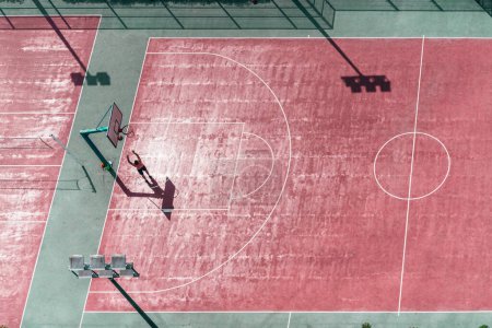 Téléchargez les photos : A drone view of a colorful basketball court with a silhouette of a man throwing the ball into the basket; view from the high above of red and green basketball playground field on a sunny day - en image libre de droit