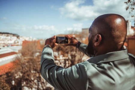 Téléchargez les photos : View from behind of a bald bearded black tourist man shooting a panorama on his smartphone from a city observation point; an African guy photographing Lisbon attraction on his cellphone - en image libre de droit