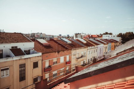 Foto de View from the rooftop of a Lisbon residential district with a narrow street full of colorful dwelling houses with clay-tiled rooftops and a cityscape in the background on a sunny day, Portugal - Imagen libre de derechos