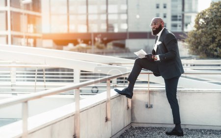 Photo for View of a fancy mature bald black businessman in a tailored formal suit and eyeglasses putting one foot on the ledge so that it would be convenient to sign the document that he holds in his hands - Royalty Free Image