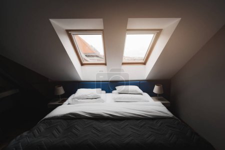 Foto de A wide-angle indoor shot of a bedroom on the last floor apartment with two windows; a hotel room in a penthouse with a made bed with a black blanket under the roof windows - Imagen libre de derechos