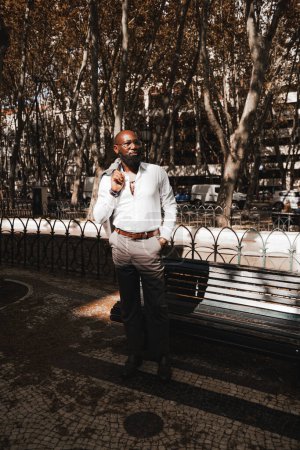 Photo for A vertical shot of an elegant mature bald African American man in a white shirt and eyeglasses, with a well-groomed black beard, standing next to a park bench, holding a jacket slung over his shoulder - Royalty Free Image