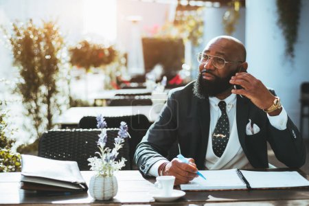 Foto de A portrait of an elegant bald bearded black man entrepreneur in a formal suit and eyeglasses in a street cafe with a notebook and a cup of coffee having a phone call; a copy space area on the left - Imagen libre de derechos