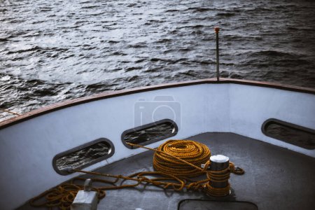 Téléchargez les photos : A low-key evening shot with a selective focus on a bollard as a chromed short and thick post on the deck of a ship to which secured an orange rope, with a luxury yacht, waves in a defocused background - en image libre de droit
