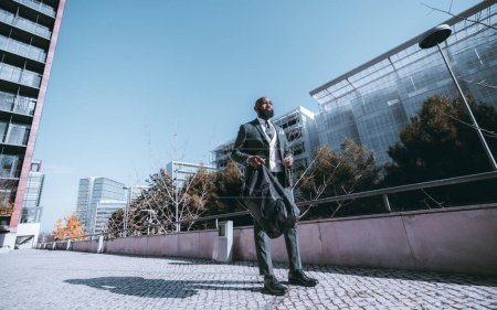 Téléchargez les photos : An elegant African executive male standing on the sidewalk, dressed in a dark grey full suit, and a white vest, and holding in one hand eyeglasses and on the other black weekender bag on a clear day - en image libre de droit