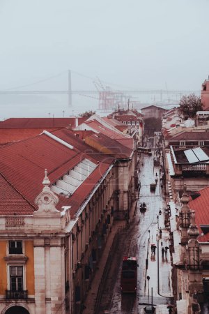 Foto de A vertical dull view of Lisbon, the 25th of April Bridge half hidden by fog in the background, the red brick roofing tiles of the buildings, and the traffic in the downtown district in wintertime - Imagen libre de derechos